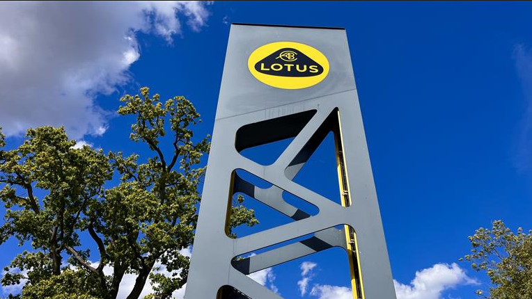 A tower-like metal sign which has the yellow and black Lotus logo at the top. It depicts a yellow circle, black triangle and the words Lotus in yellow. Beyond is a blue sky and the tops of trees in leaf.