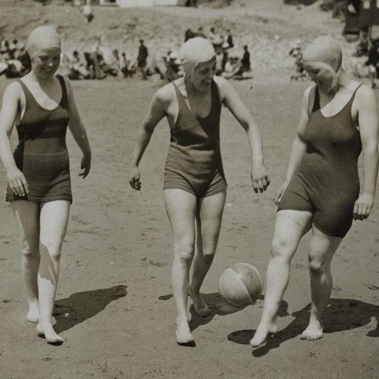 Three ladies on Porthcawl beach kicking a ball. They are wearing swimming caps and vantage-style swimsuits. 