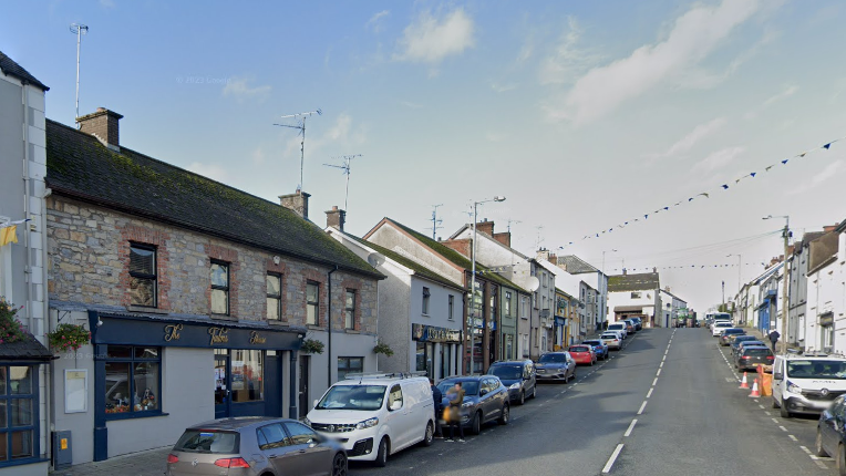 A screen grab of a Google Maps view of Main Street, Ballygawley.  The street is lined on both sides with houses, businesses and parked vehicles.  The Tailor's House pub, a two-storey building with a brick and grey rendered exterior, is in the foreground.  