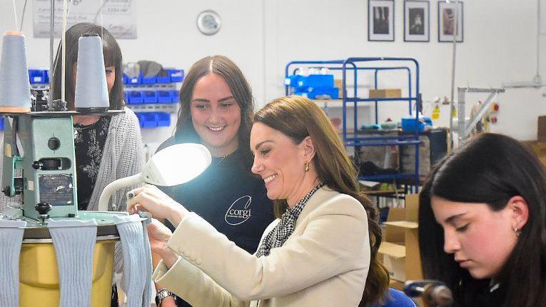 The Princess of Wales smiles as she spends time with members of the production team on the factory floor during a visit to Corgi in Ammanford. She is sitting at a workstation where goods are produced