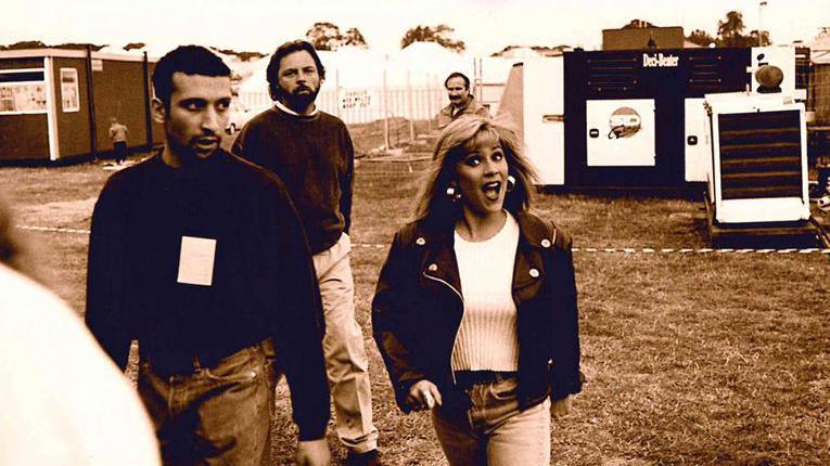 Sam Fox with long blonde hair with a surprised expression, wearing a black jacket with metal studs and a white top, with jeans. She is alongside two men, one of whom has a security pass around his neck. They are walking across a field at a festival with a hut, metal fencing and storage units behind them.