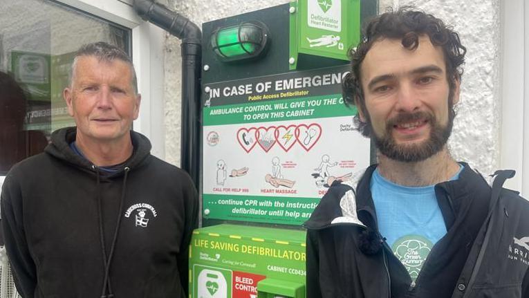 An older man in a black hooded top stands to the left of a wall mounted defibrillator. David Starley is younger and has black hair and a black beard. There is a picture besides the green defibrillator showing people how to apply it. Writing said 'ambulance control will give you the code to open this cabinet'.