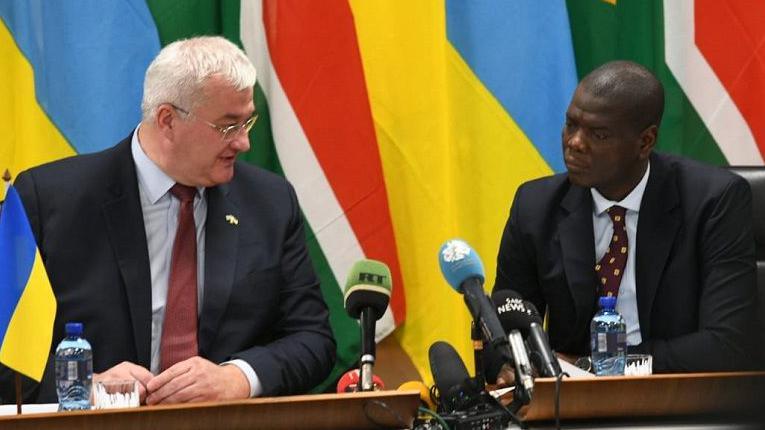 International Relations Minister Ronald Lamola on the right and his Ukrainian counterpart Andrii Sybiha sitting behind some microphones and in front of Ukrainian and South African flags as they address a press conference.