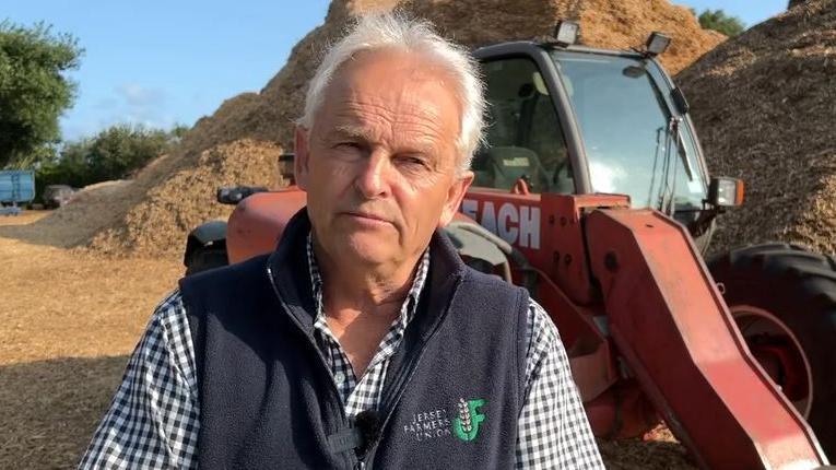 A man with short light grey hair looking just past the camera. He is wearing a black and white gingham style shirt with a dark blue fleece gilet with the Jersey Farmers Union logo on the left breast. There is a pile of hay behind him with a farming vehicle in front of that.
