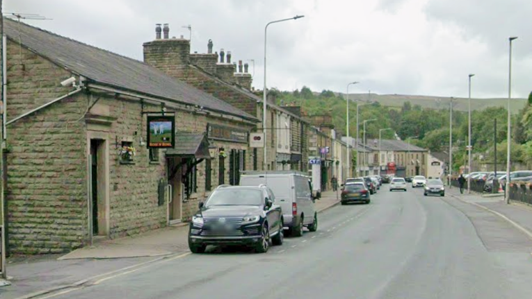 Google streetview showing old stonebuilt two-storey houses or properties lining the street with cars parked in front. Pennine hills and trees are seen in the backdrop.