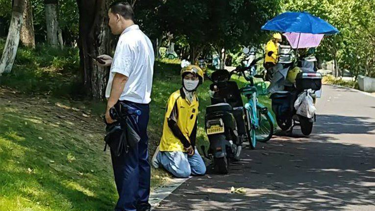 A delivery rider in a yellow uniform kneels next to their bike on the side of a road, as a security guard in a white shirt stands next to them.