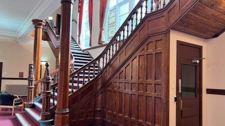 An oak-panelled staircase from the view of a hallway, which features a red carpet and large windows. The room is well-lit.