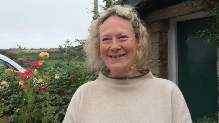 A women with sort curly blonde hair looking smiling and looking just past the camera. She is wearing a light jumper with a black collar. There is a hedge with some flowers over her left hand side shoulder and a black garage door over her right.