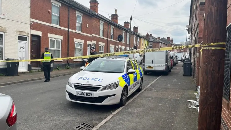 A police car is pictured with two officers, one in a hi-viz jacket, next to it. They are behind crime scene tape, which blocks off the street.