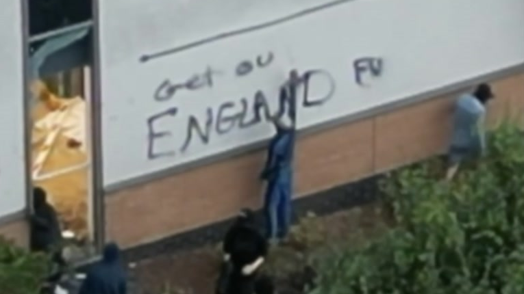 A man spray paints a grey external wall of a hotel in Tamworth with black paint. He has written 'Get out of England' and a few other men with faces covered stand nearby.