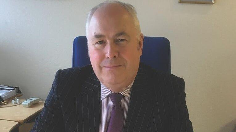 Malcolm Burr - a man with grey hair pictured sitting at his desk smiling wearing a pinstripe suit and purple tie