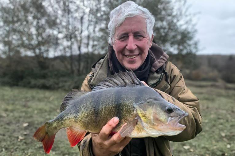 A man with grey hair in a big coat holding up a medium-sized perch fish. He is stood in a field with trees behind him.