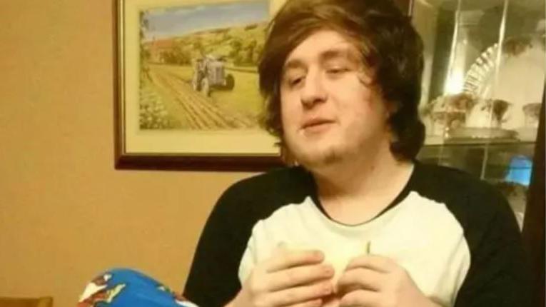 Alexander McCartney sitting in a dining room, he is wearing pyjamas. He has long dark, fuzzy hair. There is a picture of a countryside scene with a tractor in the background. He is eating a sandwhich.