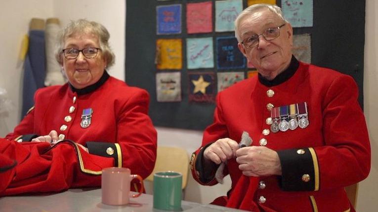 Keith Pendleton, wearing a smart red jacket with shiny buttons down the front and medals on his chest, sits alongside Anne Lloyd, who is wearing the same uniform