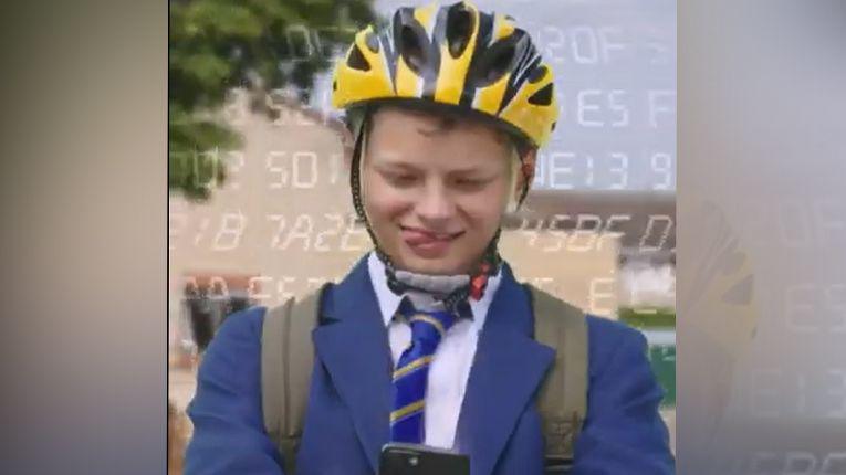 A boy in a blue blazer and striped tie has a yellow helmet on and a rucksack, and he is looking down at a phone. He is sticking his tongue out to one side of his mouth. He is outside.