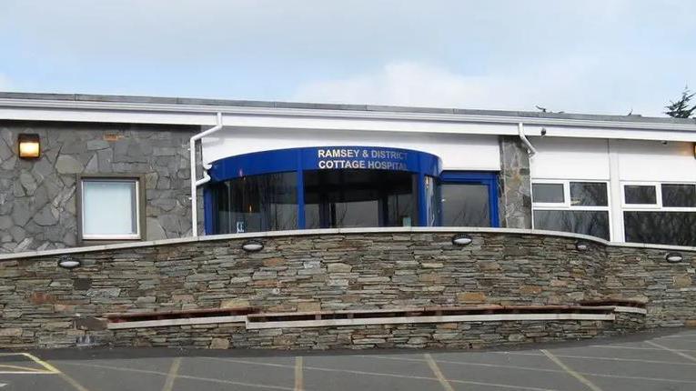 The front entrance to Ramsey Cottage Hospital, which has a blue revolving door with a stone wall in front of it and a car park.