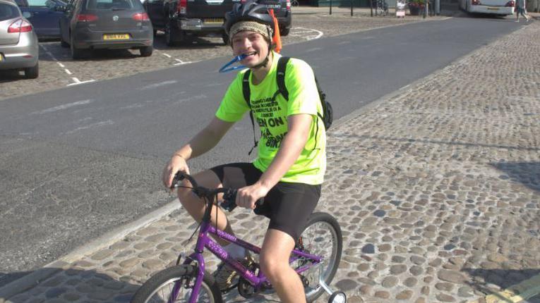 Ben Reid on a small child's purple smiling as he reaches Newcastle. He is wearing a helmet, lycra shorts and a green top