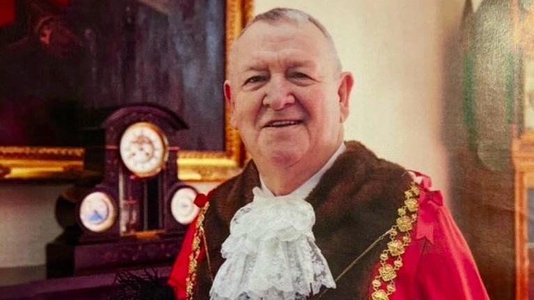 Jeff Lovell wearing his red mayoral robes with fake fur trim and a white lace jabot. He is wearing gold mayoral chains and is smiling at the camera. HE is standing in front of a fireplace with a mantle clock and a large gold framed painting above it. 