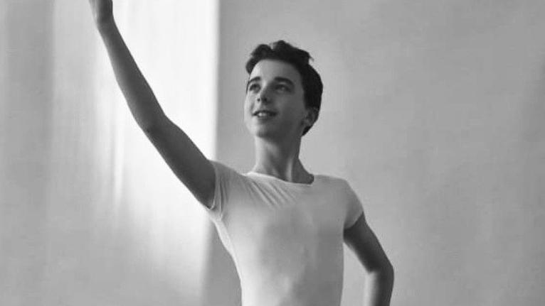 Barnaby is captured in a black-and-white photo wearing a fitted short-sleeve top. One arm is raised upward with the hand extended, while the other arm rests on the hip. The background is plain and softly lit, suggesting an indoor setting with natural light coming from the left side.