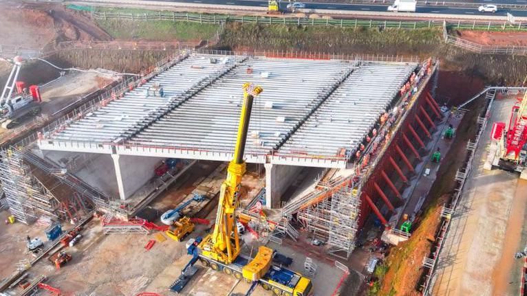 A large box-like structure with beams or girders on top sits on a building site next to a road. A large yellow crane stands next to it.