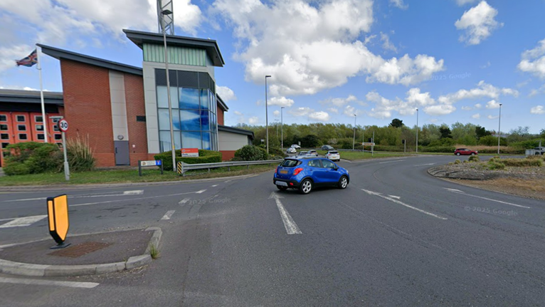 A Google Streetview image of the roundabout junction with a small blue car on the carriageway and a fire station building in the background