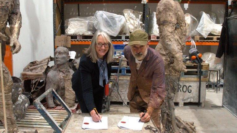 Caroline Topping and Laurence Edwards pictured in his foundry. They lean over a table, each with a white paper contract in front of them, and they both have a pen in their hand. They look at the camera and smile. Caroline has long grey hair and wears a black top and trousers. Laurence wears a green hat and a purple shirt.