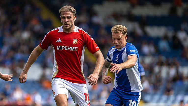 Harry Smith runs with the ball at his feet as he looks straight ahead and an Oldham player runs behind him on his left, during Swindon's game with Oldham in August 2025