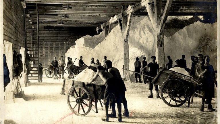 A black and white photo shows workers at a salt mine in Winsford. There are a number of workers pushing large barrows filled with salt and giant piles of sale behind them.