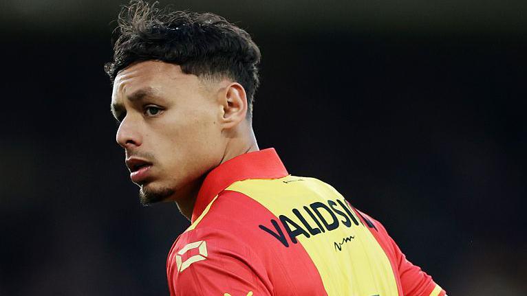 Dean James, with short dark hair and a small goatee and wearing a red and yellow striped Go Ahead Eagles shirt with black text across the back, looks over his left shoulder