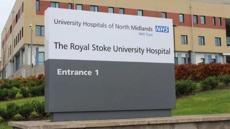 A white and grey sign outside The Royal Stoke University Hospital, with the hospital building visible behind it.