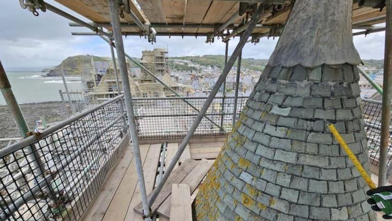 View from a scaffold looking on a slate spire down at the building. A hand with a yellow tape measure is in the lower right corner 