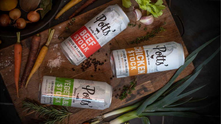 Three cans of stock, labelled beef stock, chicken stock and vegetable stock. The cans are placed on cardboard next amongst vegetables and herbs.