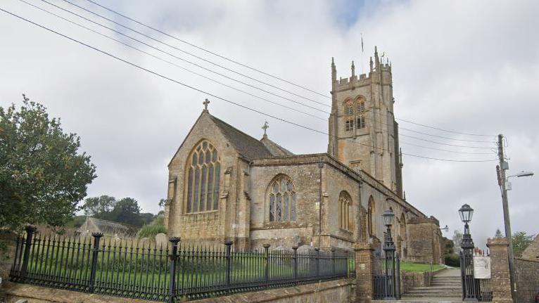 A Google Maps image of a large church with a wide tower and big windows, set among a graveyard walled off with an iron fence, and with steps leading up to it from the street.