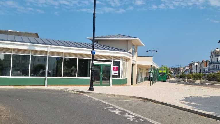 A one-storey building with lots of windows. In front of the building is a loading bay. In the background there is a green bus pulled up next to the building.