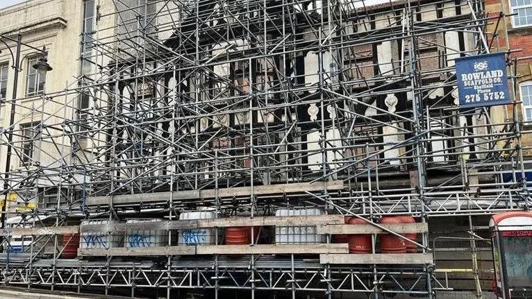 A mass of scaffolding obscures an historic black and white coloured building frontage across three floors.