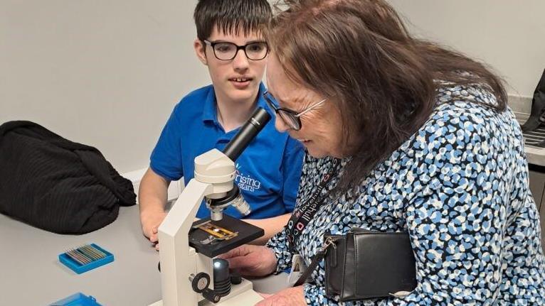 A woman with patterned dress, shoulder length brown hair and glasses looking through a microscope with a boy, who is wearing a blue polo shirt, short brown hair and glasses.
