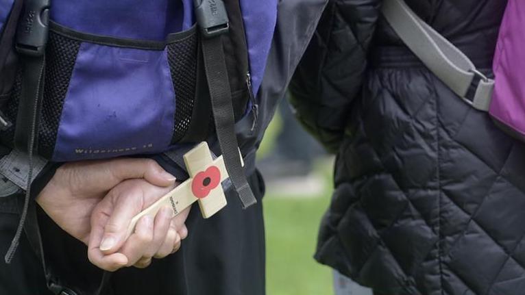 The backs of two people. Both are wearing coats and purple back packs. One of them, the one on the right, is standing with their hands behind their back. They are holding a small wooden cross with a red poppy on it.