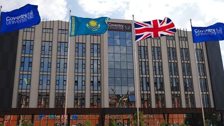 A large grey building with dozens of windows. Two flags with the Coventry University logo are placed either side of a Kazakhstan and Union Jack flag.