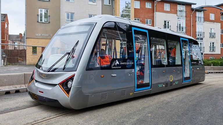 A very light railway carriage in Coventry, being run as part of a trial of a scheme. There are a number of people on board wearing orange high visibility jackets.