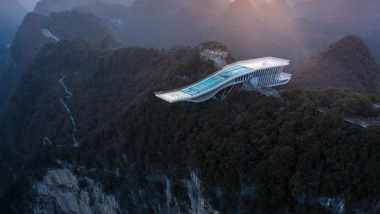 A futuristic platform reaches out from a forested cliff edge over a massive sinkhole. Mountains lit by a low sun stretch out in the background