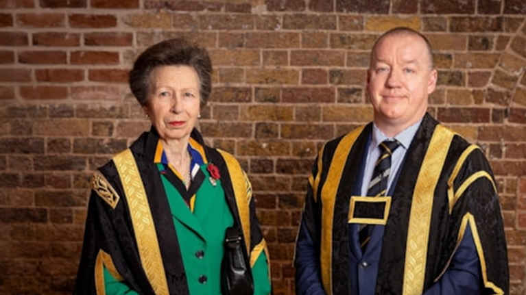 Princess Anne in black and gold robes and a green jacket stood next to a man wearing a black and gold robes and a blue suit, standing in front of a brick wall