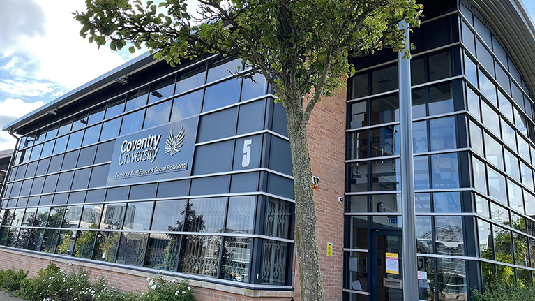 A side-view of a building at Coventry University - it has glass windows and brick walls, with the number five on the side as well as Coventry University in yellow writing. There is a tree in the foreground.