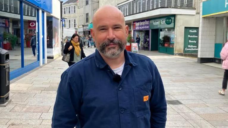 John Brown is a bald man with a dark grey beard, and is wearing a white T-shirt and navy blue overshirt. He is looking at the camera, smiling and has shops on either side of him while standing in Fore Street, St Austell.