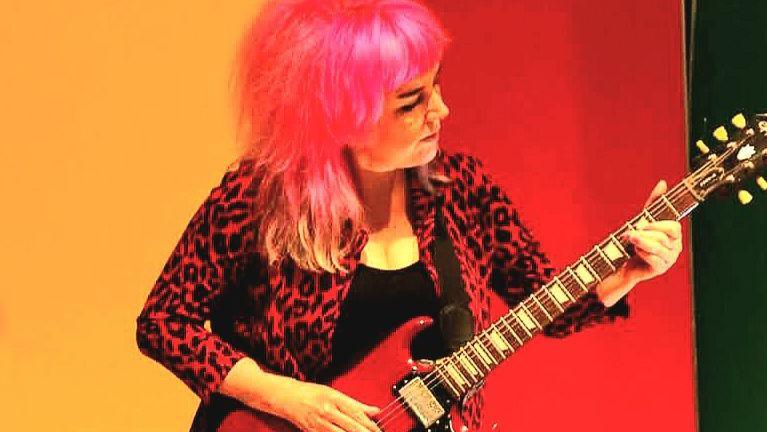 A woman with pink hair plays guitar. She is wearing a red leopard print jacket and a black T shirt.