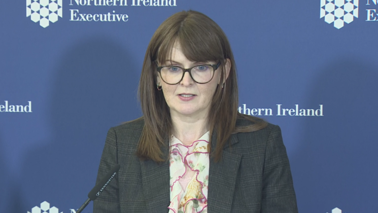 The finance minister wearing a dark grey suit and floral shirt. She stands infront of a blue board with the Northern Ireland executive logo. 