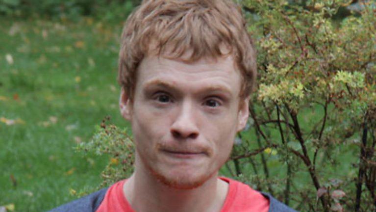 Ginger haired man looks to camera. He seems to be staring. He is wearing a red T-shirt with a blue top over it. There is a bush and green grass behind him.