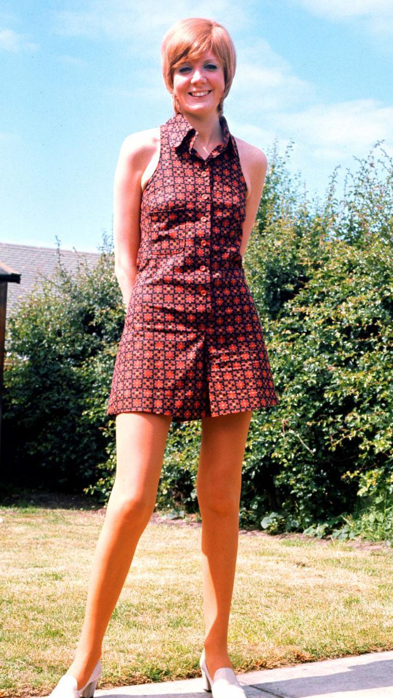 A young Cilla Black wears a short red-patterned dress with white shoes. She has short ginger hair and stands in a sun-scorched garden.