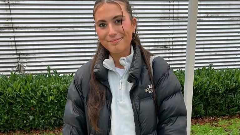 Eleanor is smiling at the camera, she has very long brown hair, tied away from her face. She is wearing a black coat and white fleece underneath. 