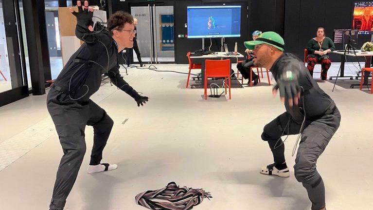 Two men face each other in a studio space wearing motion sensitive equipment, designed to record their movements