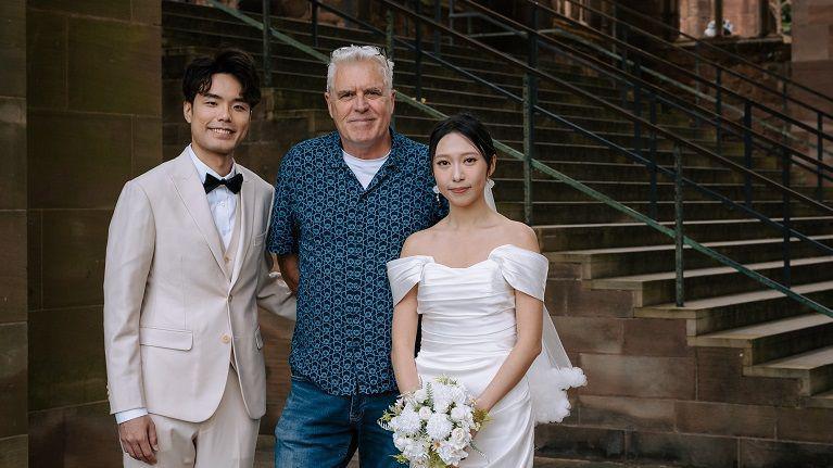 A man and woman who are from Hong Kong, standing in front of Coventry cathedral. The woman is wearing a white wedding dress and veil. The man is wearing a cream suit and black bow tie. They are posing for engagement pictures.